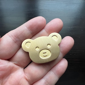 Pode incluir: Um amuleto de urso de metal dourado com um design simples e de desenho animado. O urso tem uma cara redonda com dois olhos pequenos, um nariz e uma boca.