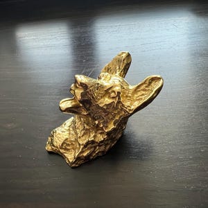 May include: A gold-colored metal sculpture of a dog's head with its mouth open, as if barking. The sculpture is detailed with textured fur and a realistic expression.