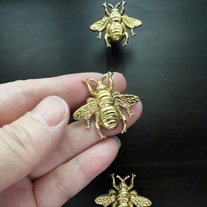 Peut inclure: Trois ornements en métal en forme d'abeille de couleur dorée. Les abeilles sont détaillées avec des lignes en relief et ont une finition brillante.