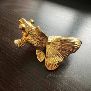 May include: A gold-coloured metal sculpture of a fish with a detailed, textured surface. The fish is positioned with its tail raised and its head tilted upwards.