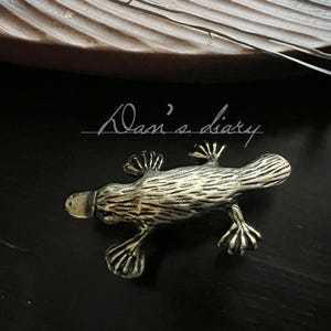 May include: A detailed, metallic platypus figurine with textured details, including webbed feet and a duck-like bill. The text "Dan's diary" is written in cursive above the figurine. The figurine is set against a dark background.