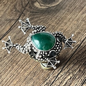 Può includere: Spilla a forma di rana color argento con una grande gemma verde a forma di goccia al centro. La spilla presenta dettagli strutturati ed è appoggiata su una superficie di legno.