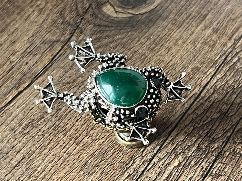 以下が含まれることがあります： 銀色のカエル型ブローチ。中央に大きな涙型の緑色の宝石が配置されています。ブローチは詳細なテクスチャ加工が施されており、木製の表面に置かれています。