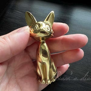 Könnte beinhalten: Eine goldfarbene Metallfigur eines Katers mit großen, runden Augen. Der Kater sitzt aufrecht mit dem Schwanz um seinen Körper gewickelt.