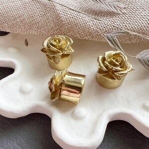 May include: Three gold-colored bottle stoppers with rose designs. The stoppers are on a white, textured surface.