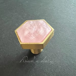 May include: A hexagonal cabinet knob with a pink resin insert and a gold metal base.