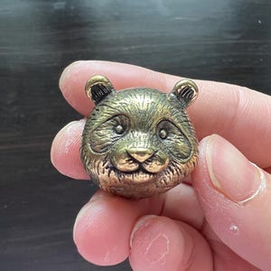 Puede incluir: Una detallada escultura de cabeza de panda de color bronce. La cara del panda está intrincadamente diseñada con ojos, nariz y orejas visibles. La escultura tiene una apariencia vintage o antigua, lo que sugiere que podría ser un artículo decorativo o de colección.