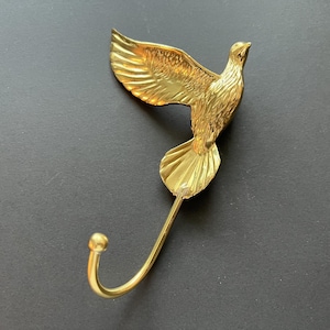 Op de afbeelding: Een gouden metalen haak in de vorm van een duif met een gebogen haak aan de onderkant.