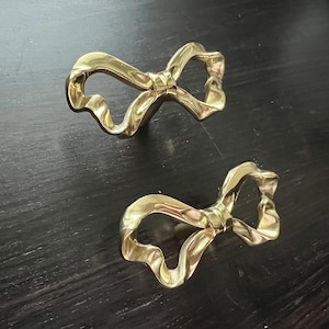 May include: Two gold-colored bow-shaped cabinet pulls. The pulls have a twisted ribbon design and are set against a dark wood surface. These decorative hardware pieces add a touch of elegance.