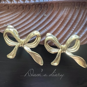 May include: A pair of gold-colored bow-shaped earrings. The earrings are made of a shiny metal and are resting on a dark surface. The earrings are in a bow shape with detailed curves and folds. The text "Dani's diary" is visible.
