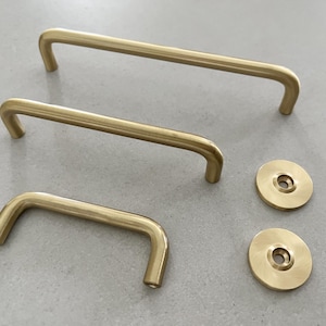 Peut inclure: Trois poignées de meubles en laiton et deux plaques arrière rondes assorties. Les poignées sont incurvées, avec une finition lisse et mate. La quincaillerie est disposée sur une surface gris clair, mettant en valeur un design minimaliste.