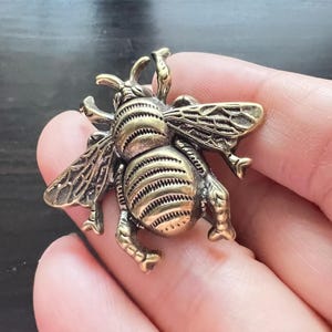 Op de afbeelding: Een gedetailleerde, bronskleurige bijenhangers. De bij heeft ingewikkelde vleugel- en lichaamsdetails, met een getextureerd oppervlak. De vleugels zijn uitgespreid en de bij staat in een levensechte pose.