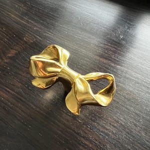 May include: A gold-coloured metal bow tie shaped like a ribbon. The bow tie is a decorative object.