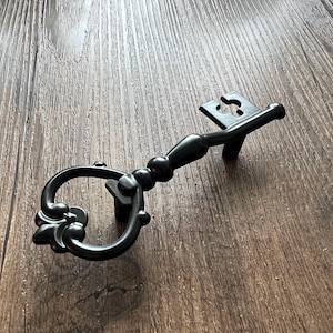 Può includere: Pomello per armadio decorativo nero a forma di chiave con maniglia decorativa e design a buco della serratura. La chiave è in metallo ed è posta su uno sfondo a venature di legno. Questo hardware in stile vintage aggiunge un tocco unico ai mobili.