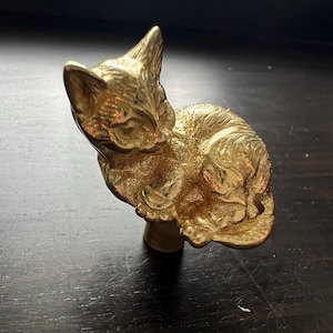 Puede incluir: Una figurita de metal dorada de un gato durmiendo. El gato está enroscado en una pequeña base.