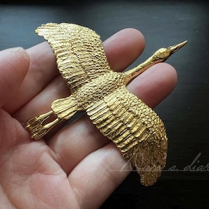 Könnte beinhalten: Eine goldfarbene Metall-Brosche in Form eines Kranichs mit ausgebreiteten Flügeln. Die Brosche ist mit strukturierten Federn verziert und wird in einer Hand gehalten.