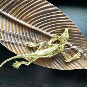 May include: A gold-colored lizard figurine on a wooden leaf-shaped tray. The lizard is posed in a curled position with its tail wrapped around the tray.