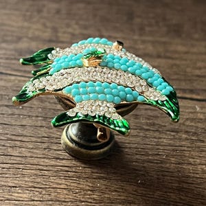 以下が含まれることがあります： 魚の形をした装飾的なキャビネットノブ。魚はターコイズと白で、クリスタルのアクセントと緑色のひれが付いています。ノブはブロンズ色のベースに取り付けられています。