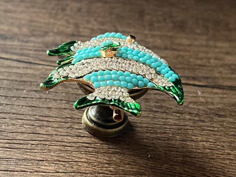 以下が含まれることがあります： 魚の形をした装飾的なキャビネットノブ。魚はターコイズと白で、クリスタルのアクセントと緑色のひれが付いています。ノブはブロンズ色のベースに取り付けられています。