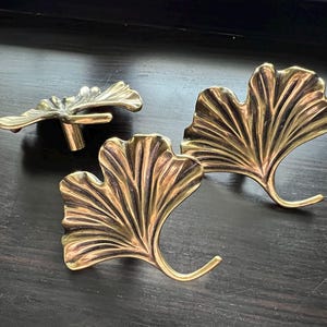May include: Three brass-colored Ginkgo leaf-shaped cabinet hardware pieces. The set includes two pulls and a knob, all with detailed veining. The hardware is designed to be mounted on a cabinet or drawer, adding a decorative touch.