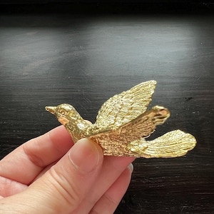 Może przedstawiać: Złota metalowa figurka gołębia z rozpostartymi skrzydłami. Gołąb jest w locie i ma szczegółową, fakturowaną powierzchnię.