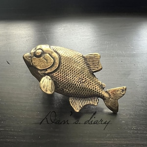 May include: A detailed, brass-colored fish figurine with textured scales, fins, and tail. The fish is positioned on a dark surface, with the handwritten text "Dan's diary" visible below it. The figurine has a vintage aesthetic.