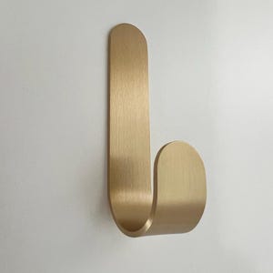 Może przedstawiać: Złoty metalowy haczyk o zakrzywionym kształcie. Haczyk jest zamontowany na białej powierzchni. Haczyk ma prostokątny wierzch i zakrzywione dno, odpowiedni do wieszania przedmiotów.