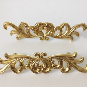 May include: Two ornate gold-toned cabinet pulls with a decorative, scroll-like design. The pulls are made of metal and have a shiny finish.