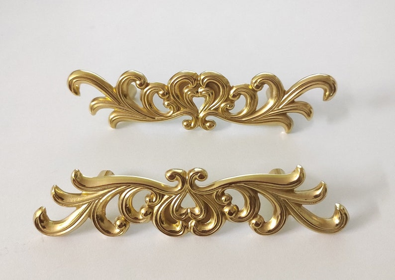 May include: Two ornate gold-toned cabinet pulls with a decorative, scroll-like design. The pulls are made of metal and have a shiny finish.