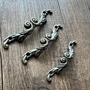 May include: Three antique-style metal decorative pulls with an ornate, swirling leaf design. Each pull has two holes for mounting. The pulls are a muted silver color and rest on a wooden surface.