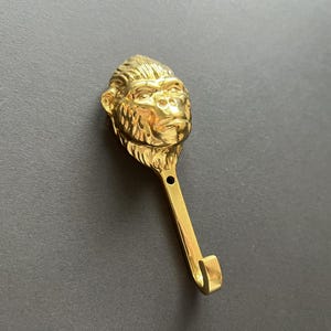 Könnte beinhalten: Ein goldfarbener Metallhaken in Form eines Gorillakopfes. Der Haken ist zum Aufhängen von Gegenständen an einer Wand gedacht.