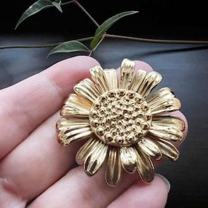 May include: A gold-toned metal brooch in the shape of a sunflower. The sunflower has a textured centre and petals.