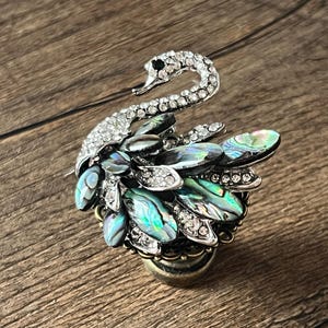 Può includere: Spilla a forma di cigno dettagliata con corpo e testa argentati, incastonata con strass trasparenti. Le ali presentano dettagli iridescenti in conchiglia di abalone e strass aggiuntivi. La spilla è montata su una base dorata.