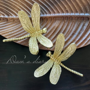 May include: Two gold-coloured dragonfly-shaped decorative objects. The dragonflies have detailed wings and bodies, and are placed on a wooden surface with a leaf-like design. The background is dark.