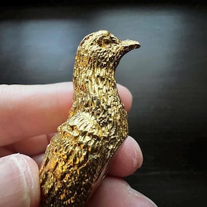 Może przedstawiać: Szczegółowa, złota figurka ptaka z teksturowanymi piórami i zakrzywionym dziobem. Figurka jest trzymana w dłoni, prezentując skomplikowany design i metaliczne wykończenie. Głowa ptaka jest obrócona na bok.