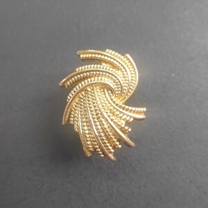 K&ouml;nnte beinhalten: Eine goldfarbene Vintage-Brosche mit einem wirbelnden, strukturierten Design. Die Brosche hat ein Spiralmuster mit detaillierten, geriffelten Kanten, wodurch ein dreidimensionaler Effekt entsteht. Das Schmuckst&uuml;ck ist vor einem dunkelgrauen Hintergrund platziert.