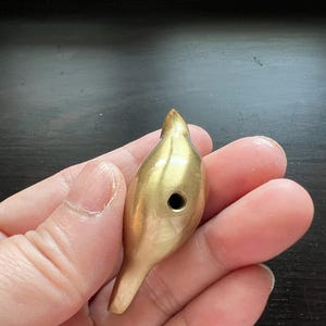 Pu&ograve; includere: Un fischietto per uccelli in metallo dorato con un piccolo foro al centro. Il fischietto ha la forma di un uccello con un becco appuntito e un corpo arrotondato.