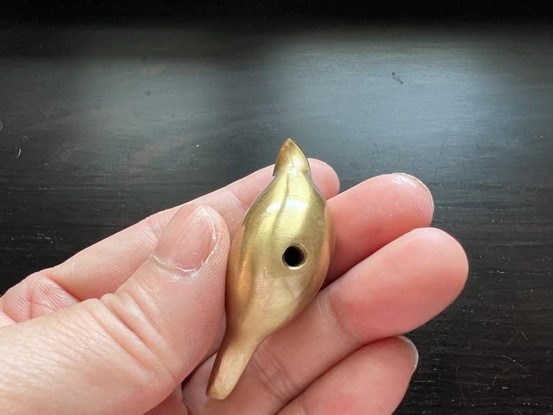 Pu&ograve; includere: Un fischietto per uccelli in metallo dorato con un piccolo foro al centro. Il fischietto ha la forma di un uccello con un becco appuntito e un corpo arrotondato.