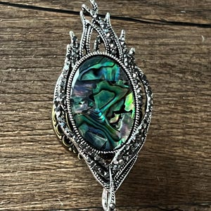 May include: An elaborate, silver pendant featuring an oval, iridescent abalone shell. The pendant has a decorative, flame-like design with small, dark crystals. The clasp is a silver bail.