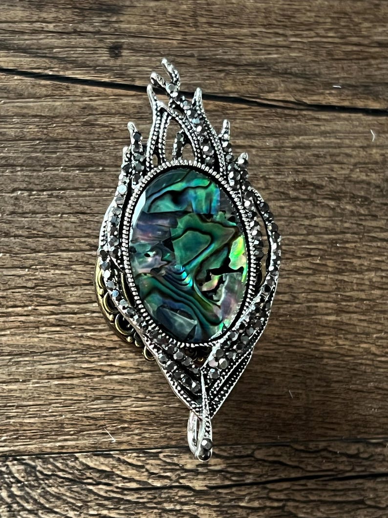May include: An elaborate, silver pendant featuring an oval, iridescent abalone shell. The pendant has a decorative, flame-like design with small, dark crystals. The clasp is a silver bail.