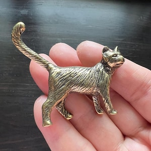 Op de afbeelding: Gedetailleerd messing kattenbeeldje met een getextureerd oppervlak. De kat staat met zijn staart omhoog gekruld. Het beeldje heeft een vintage esthetiek en is waarschijnlijk een decoratief item.