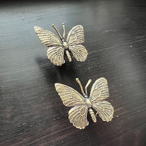May include: Two antique brass butterfly cabinet knobs. The knobs feature detailed wing patterns and antennae. The butterflies are set against a dark wooden surface, creating a contrast in colour and texture. These knobs are ideal for adding a touch of nature to furniture.