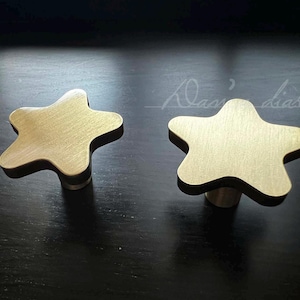 Può includere: Due pomelli per mobili a forma di stella color oro. I pomelli hanno una finitura spazzolata e sono montati su una superficie nera.