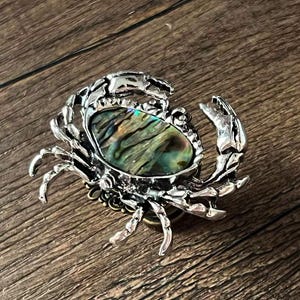 Può includere: Spilla a forma di granchio color argento con un centro ovale in conchiglia di abalone. Il guscio mostra tonalità iridescenti verdi, blu e oro. La spilla ha artigli e zampe dettagliati, su una superficie di legno.