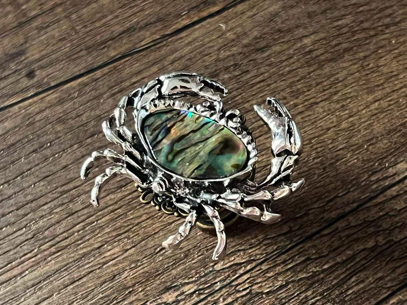 K&ouml;nnte beinhalten: Silberfarbene Krabbenbrosche mit ovalem Abalone-Muschel-Mittelteil. Die Muschel zeigt schillernde gr&uuml;ne, blaue und goldene Farbt&ouml;ne. Die Brosche hat detaillierte Krallen und Beine, vor einem Holzuntergrund.