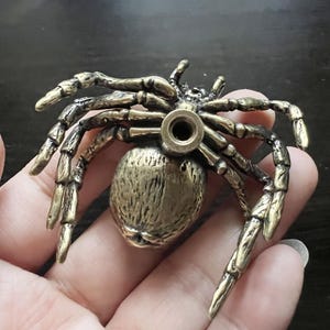 K&ouml;nnte beinhalten: Detaillierte Spinnenfigur aus Messing mit acht beweglichen Beinen und einem strukturierten K&ouml;rper. Die Spinne hat ein Loch in ihrem Hinterleib. Die Figur wird in einer Hand gehalten.