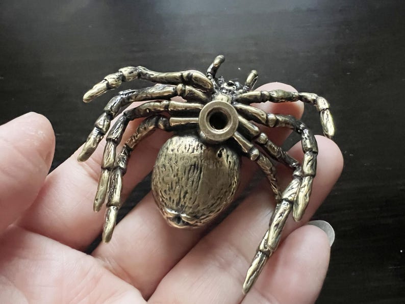K&ouml;nnte beinhalten: Detaillierte Spinnenfigur aus Messing mit acht beweglichen Beinen und einem strukturierten K&ouml;rper. Die Spinne hat ein Loch in ihrem Hinterleib. Die Figur wird in einer Hand gehalten.