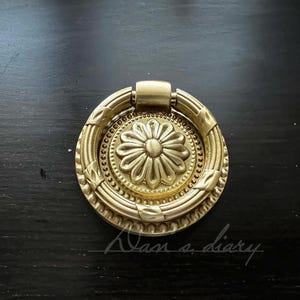 May include: A gold-toned metal ring pull with a decorative floral design. The ring pull has a smooth, polished finish and a raised floral pattern.