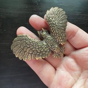 K&ouml;nnte beinhalten: Detaillierte Eulenfigur aus bronzefarbenem Metall mit ausgebreiteten Fl&uuml;geln. Die Federn der Eule sind aufwendig geschnitzt. Die Figur wird in einer Hand gehalten. Die Spannweite betr&auml;gt ca. 7,6 cm.