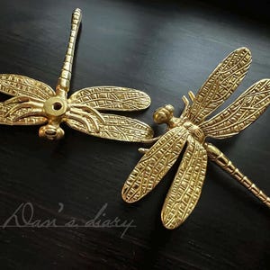 Puede incluir: Dos pomos de puerta de libélula de color dorado con intrincados detalles de alas. Los pomos son de metal y tienen un acabado pulido.